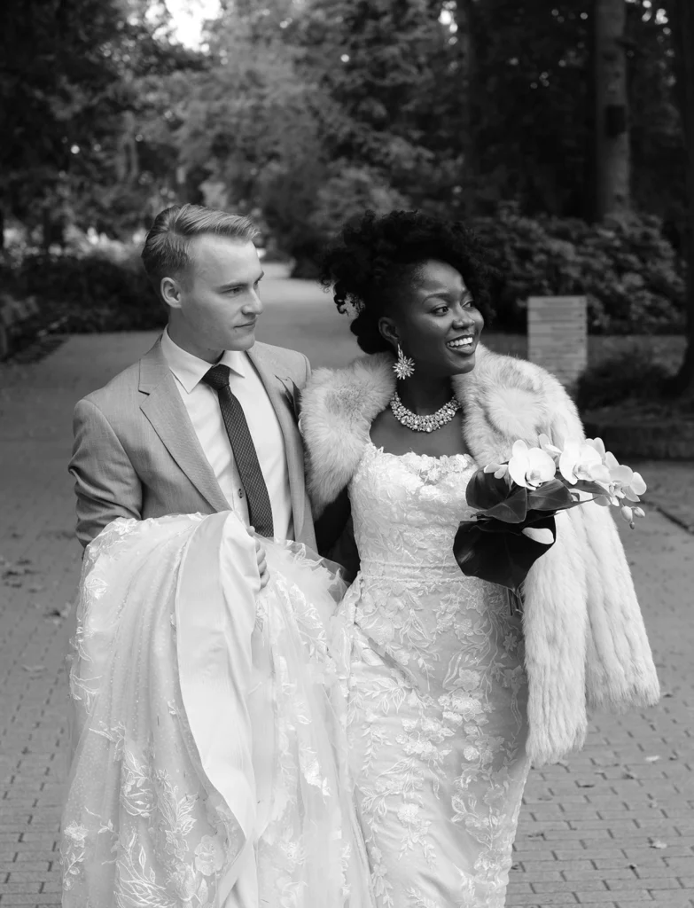 black and white image of a beautiful mixed couple is walking towards Frankfurt am Main Palmengarten she is wearing a long lace wedding dress and a fur coat